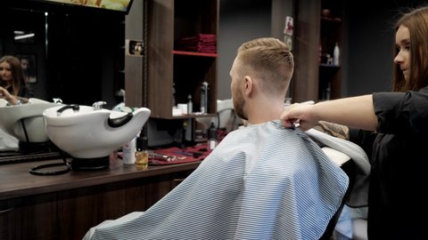 Young woman hairdresser with long blond hair covers a man in a barber chair with a cape of a client: Gomel, Belarus - July 2020