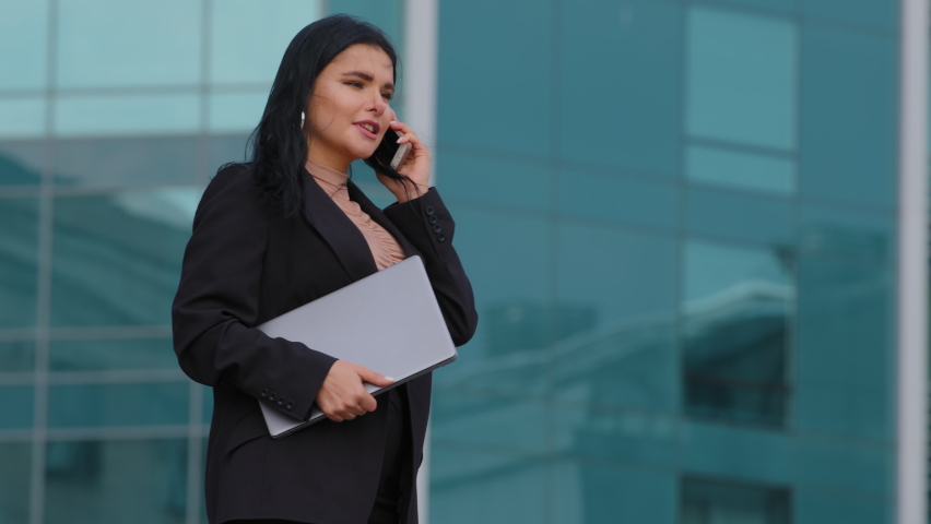 Successful young businesswoman walks down street against background of city building holds laptop confident hispanic girl manager talking by mobile phone answering business call speaking on cellphone | Shutterstock HD Video #1086510104