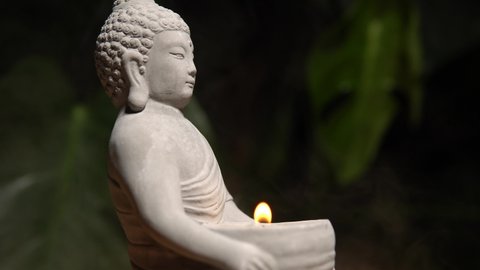Buddha made of stone on wooden table surface at natural exotic jungle green background with monstera plant. Buddha statue with burning candle fire and water steam.