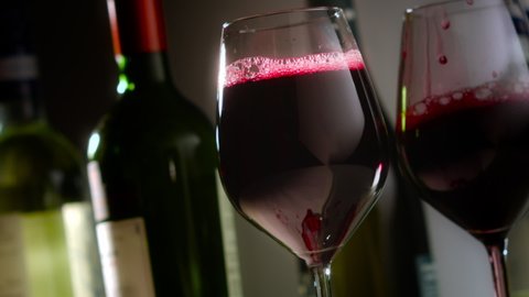 Red wine is clumsily poured into a pair of wine glasses, the wine misses and spills. Several wine bottles are in soft focus in the background.