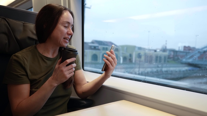 An adult woman travels by train and communicates via the Internet via video link. A lady on the train is sitting at a table with a disposable cup of coffee.