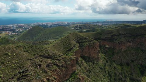 Birds eye view over the edge of a volcano crater in Gran Canaria against the backdrop of lush green valleys, small villages of Canary islands, and the Atlantic ocean in the background.