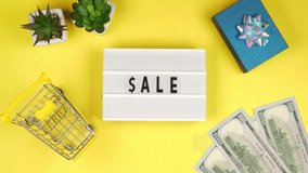 The inscription Sale on a white board  with houseplants, dollars and a toy grocery cart on a yellow background