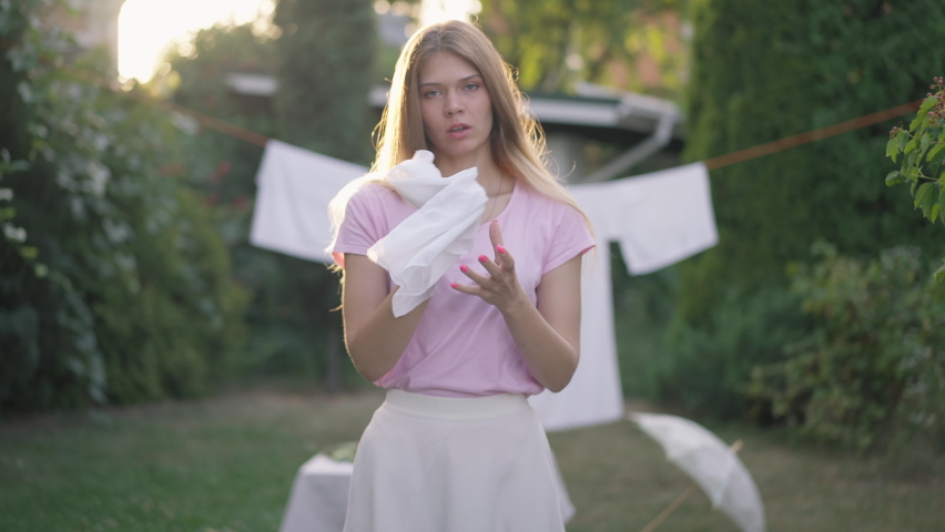Front view portrait of confident young woman rubbing hands looking at camera. Slim attractive Caucasian rural lady posing in sunshine outdoors in backyard garden with clean laundry at background