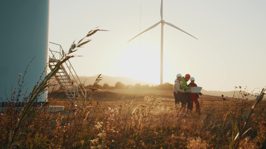 Group of engineers in hard hat walking using blueprint in windmill farm while inspecting wind turbine power station. At sunset. Slow motion