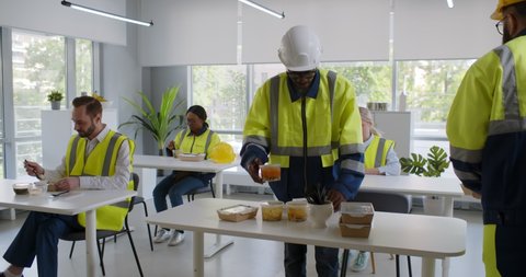 Group of diverse engineers and workers enjoy eating food and relax in canteen. Construction company employees in uniform and hardhat eating lunch in office
