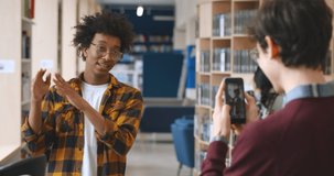Young african blogger recording vlog on digital smartphone cam in library. School student vlogger talking looking at mobile phone with friend filming him in library