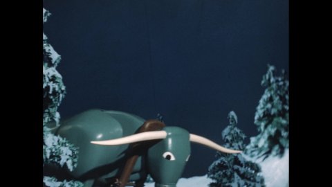 1950s: Slate. Marionette of a blue ox bows and pulls two sleds of sticks through snow covered woods. Slate. Ox pulls sled and bows. Slate.
