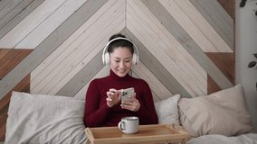 Young asian ethnic woman using smart phone surfing social media, checking news, playing mobile games or texting messages sitting on bed at home. Mixed race girl spending time with gadget technology.