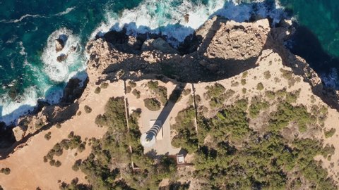 Aerial top-down view of Punta Moscarter lighthouse in Ibiza, Spain. Moving up showing the lighthouse and clear ocean and skies, waves pounding on cliffs.