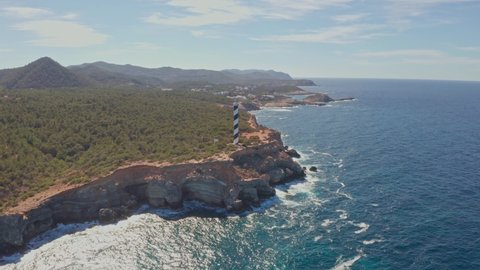 Aerial view of Punta Moscarter lighthouse in Ibiza, Spain. Panning around the lighthouse showing clear ocean and some clouds in skies, waves pounding on cliffs with village in background.