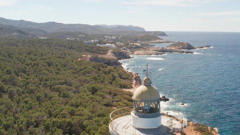 Aerial view close-up of Punta Moscarter lighthouse in Ibiza, Spain. Showing the spinning lights inside the lighthouse with clear ocean and skies, waves pounding on cliffs with village in background.