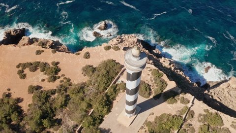 Aerial view tilting up on Punta Moscarter lighthouse in Ibiza, Spain. Showing the lighthouse close up and showing clear ocean and skies, rough waves pounding on the cliffs.