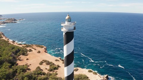 Aerial view close-up of Punta Moscarter lighthouse in Ibiza, Spain. Panning right fast, around the lighthouse showing clear ocean and skies, waves pounding on cliffs with village in background.