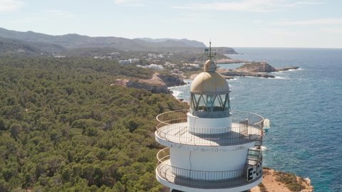 Aerial view close-up of Punta Moscarter lighthouse in Ibiza, Spain. Showing spinning lights inside the lighthouse with clear ocean and skies, waves pounding on cliffs with village in background.