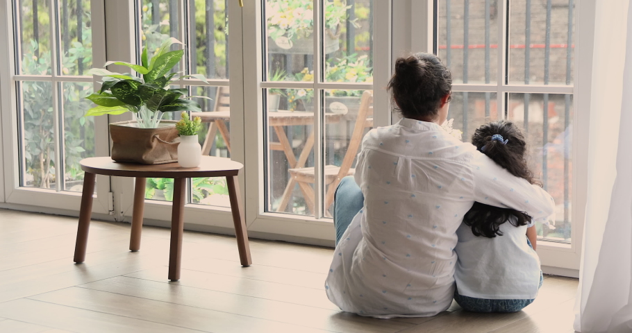 Rear back view young mom hugging her little preschool daughter sit on warm floor near window looking outside, talking, admire view from modern flat enjoy time together at home. Leisure, tenant concept Royalty-Free Stock Footage #1086555239