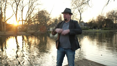 [4k] tenor with hat singing an opera at the park in front of sunset at pond