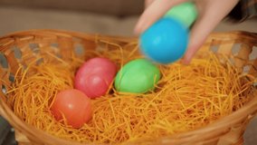 A child's hand puts Easter colored eggs in a yellow wicker basket. Holiday attributes