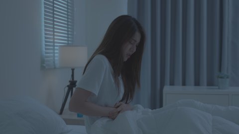 Asian beautiful girl in pajamas feel sick while sit on bed in bedroom. Attractive sleep young woman touching abdomen feeling bad with fever and stomach ache after wake up on bed in the morning at home