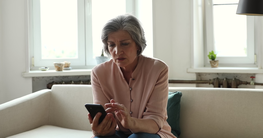 Older woman read awful sms received message, looks disappointed shocked sit on sofa with cellphone at home. Mature female staring at smartphone screen experiences device problems, need repairs concept Royalty-Free Stock Footage #1086568676