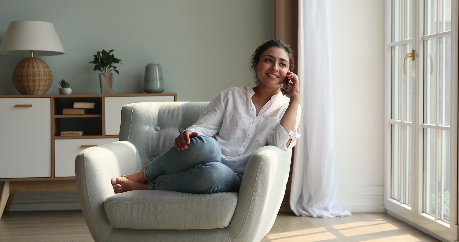 Cute Indian woman relaxing sit on armchair in cozy living room holds cellphone talking to friend, lead personal conversation on smartphone, having distancing communication. Tech, phone call concept Royalty-Free Stock Footage #1086568706