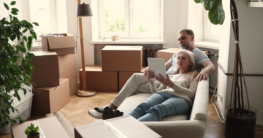 Senior couple relax on sofa in living room on relocation day near boxes, use digital tablet buy goods on internet, make order via e-service, make purchase online, search house renovation ideas concept Royalty-Free Stock Footage #1086568733
