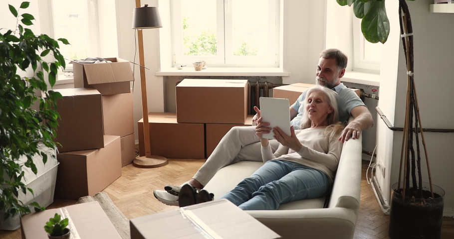 Senior couple relax on sofa in living room on relocation day near boxes, use digital tablet buy goods on internet, make order via e-service, make purchase online, search house renovation ideas concept Royalty-Free Stock Footage #1086568733