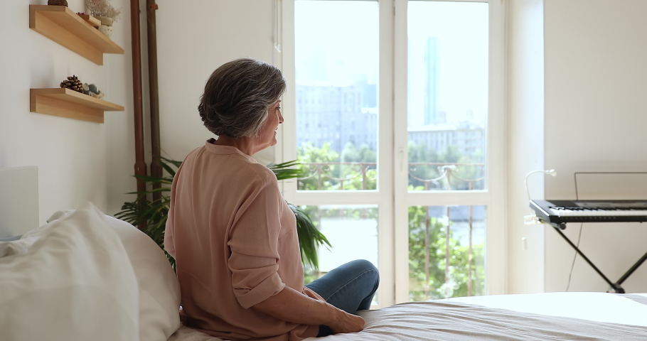 Rear view older woman in casual clothes sit on bed in modern bedroom looking out window, raise hands stretching body muscles feels happy, welcoming new day, good morning, carefree retired life concept Royalty-Free Stock Footage #1086568742
