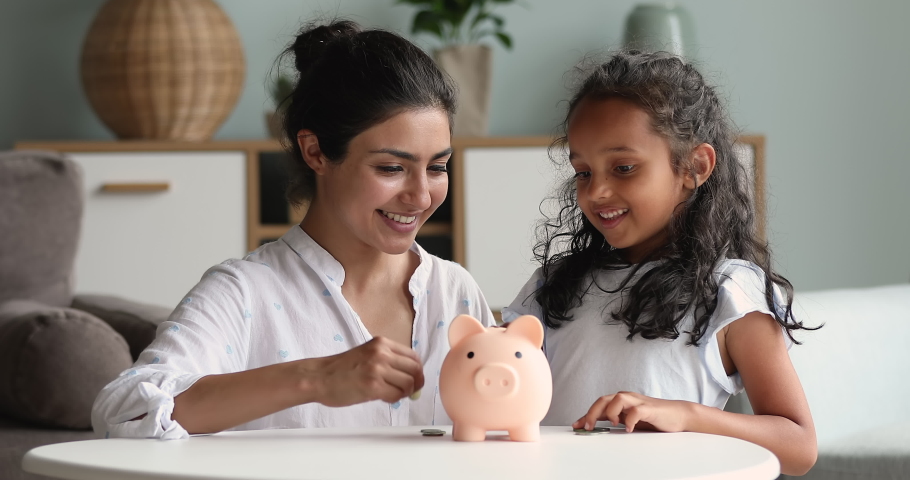 Indian mother and little daughter put coins into piggy bank give high five looking happy. Mom teach child be thrifty, save pocket money for future education or purchases, think about tomorrow concept Royalty-Free Stock Footage #1086568748