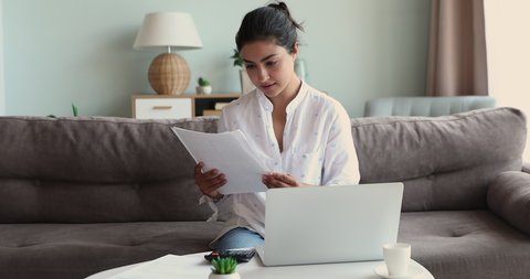 Serious Indian woman sit on sofa at table calculates expenses, pay utility bills use e-bank on laptop, sorts out paper, check documents, manage budget, control outcomes and incomes, accounting concept