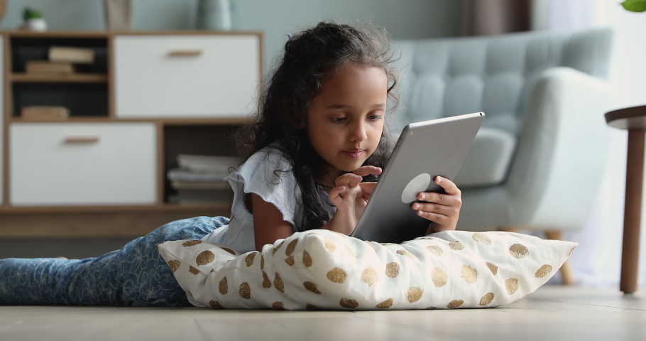 Adorable preschool Indian girl lying in living room on warm floor using digital tablet, play video games, watch favourite vlogger, spend free time alone at home. Gen Z use modern wireless tech concept Royalty-Free Stock Footage #1086568826