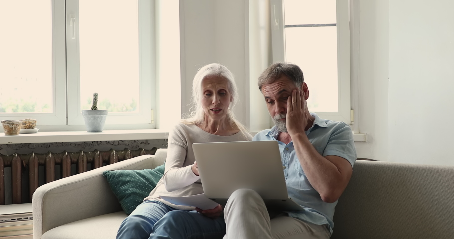 Older couple sit on sofa with laptop, reviewing expenses, learn documents from bank about debt, overspending, having financial problems feeling stressed. Bankruptcy, lack of money to pay bills concept | Shutterstock HD Video #1086568838