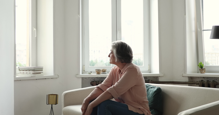 Serious older woman staring out window spend time alone at home, looking restless or disappointed, waiting missing someone seated on couch in living room. Loneliness, solitude on retirement concept Royalty-Free Stock Footage #1086568844