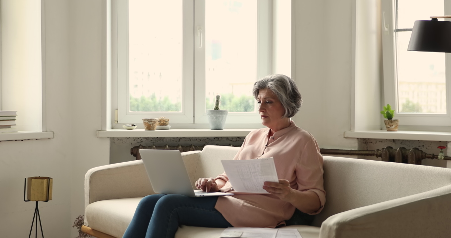 Older grey-haired woman sits on sofa with laptop pay bills through online e-bank app looking puzzled or troubled, analyze papers feels puzzled, having mistake in document, financial concerns concept Royalty-Free Stock Footage #1086568850