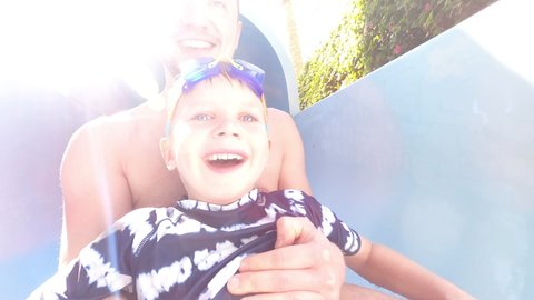 Happy smiling father rolling down the water slide with his little kid son on a clear beautiful water at Summer time