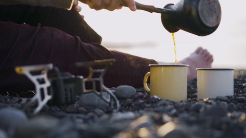 Unrecognizable barefoot woman hiker pours coffee from cezve into white cup sitting on grey pebble against endless sea under bright summer morning sunlight detail view