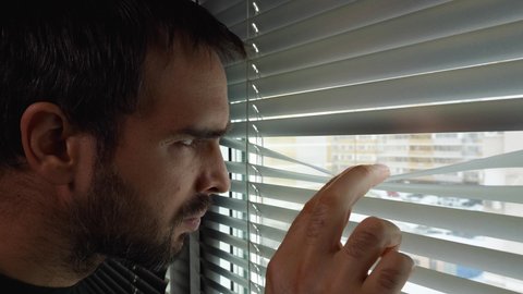 A man with a beard approaches the window, pulls back the blinds, and looks out the window through a crack. A middle-aged Caucasian man looks out the window