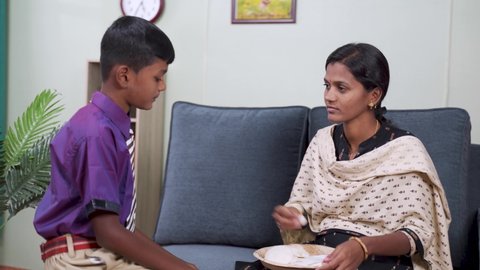 young indian mother convincing kid to eat breakfast by giving mobile phone at home - concept of bothering kid, smartphone addiction and relationship.