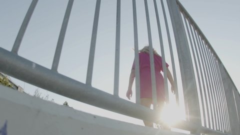 Girl in dress riding skateboard along fence outdoor. Tracking shot of female skater training in skate park on sunny summer day alone. Extreme sport, physical activity concept