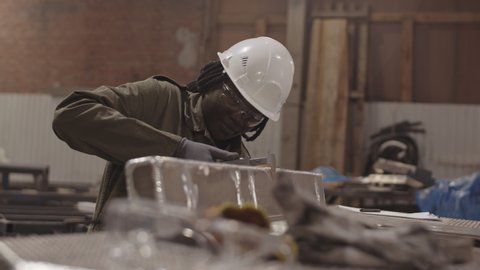 Medium close-up of female African American worker wearing white hard hat and safety goggles, using caliper measuring width of metallic piece, then reconciling dimensions on phone