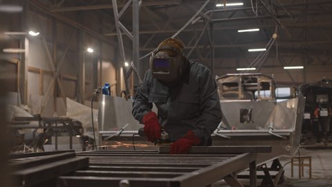 Medium shot of man wearing helmet, worker jacket and red gloves, standing in indoor dockyard, using sander on metallic pipes
