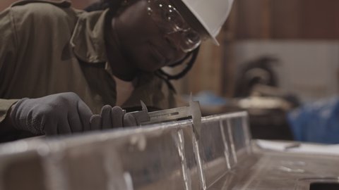 Medium close-up of female African American worker wearing white hard hat and safety goggles, leaning forward, using caliper measuring width of metallic piece