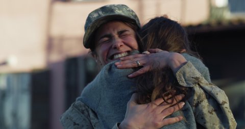 Cinematic close up shot of young female soldier mother in camouflage military uniform and cap is hugging emotionally her daughter and cries of happiness just turn back home after mission.