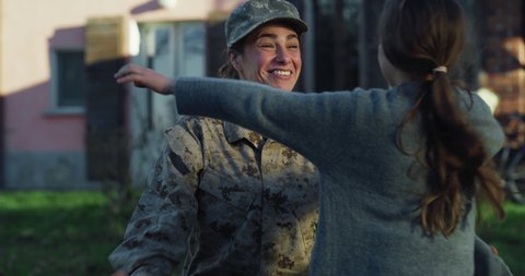 Cinematic shot of young female soldier mother in camouflage military uniform and cap is hugging emotionally her daughter and cries of happiness just turn back home after mission.