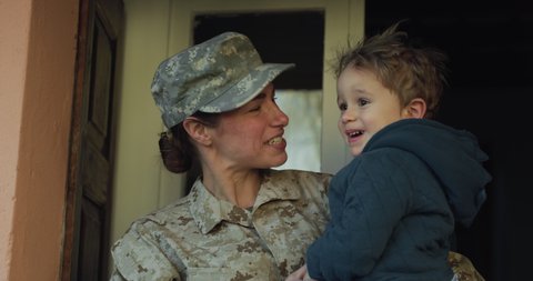 Cinematic close up shot of young happy female soldier mother in camouflage military uniform and cap is carrying and kissing with love her little toddler boy son just turn back home after mission.
