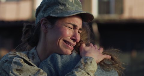 Cinematic close up shot of young female soldier mother in camouflage military uniform and cap is hugging emotionally her daughter and cries with promises to turn back before leaving home for mission.