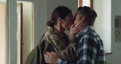 Cinematic shot of young female soldier wife in camouflage military uniform and cap is hugging emotionally her husband and cries with promises to turn back before leaving home for mission.