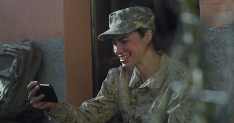 Cinematic shot of young happy smiling female soldier in camouflage military uniform and cap is making technology video call to her family or friends just turn back home after mission.