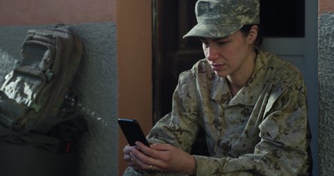 Cinematic shot of young happy smiling female soldier in camouflage military uniform and cap is technology smartphone for work or entertainment just turn back home after mission.
