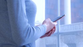 Lonely young woman browsing internet news feed on mobile phone near window at home. Young adult femle using modern smartphone for online entertainment and communication in 4k stock video